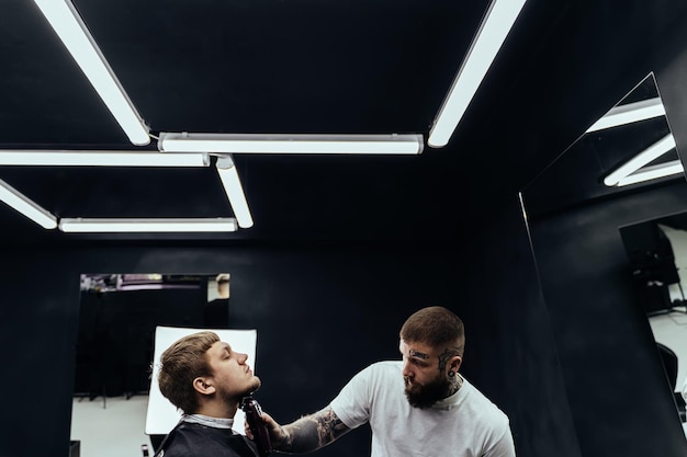 Barbeiro tatuado cortando homem barbudo com máquina de barbear em barbearia processo de penteado penteado