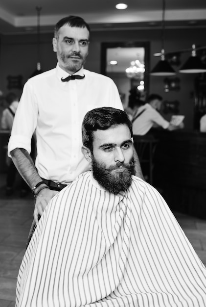 Barber hace un corte de pelo para un joven apuesto con barba y bigote en la superficie de Barbashop