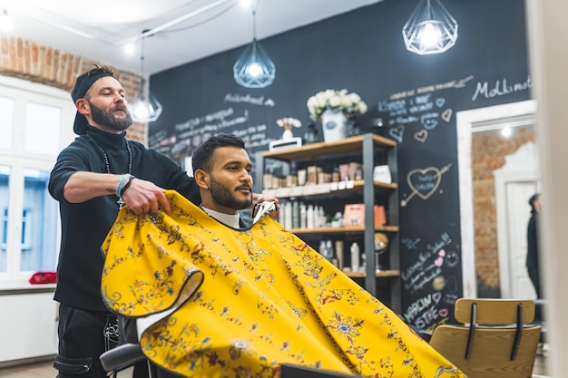 Barber poniendo una capa amarilla impresa sobre su cliente indio antes de un corte de pelo