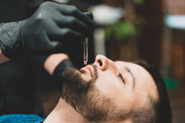 Barbería. El cliente de los maestros en la barbería, el barbero aplica aceite y cosméticos a la barba de los clientes. Tienda de belleza masculina. Estilo de vida saludable y belleza.