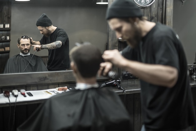 Barbero inigualable con barba y tatuaje corta el cabello de su cliente barbudo en la barbería