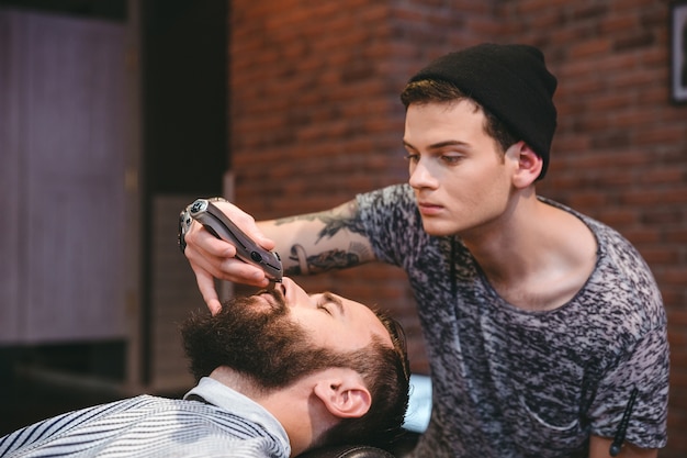 Barbero joven hábil concentrado recortando la barba de joven guapo en peluquería