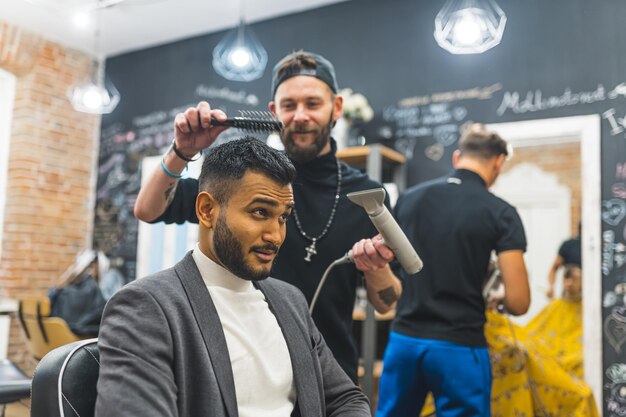 Un barbero profesional peinando el cabello de su cliente indio con una secadora en una barbería