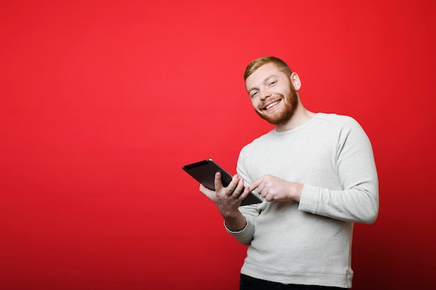 Barbudo atraente sorrindo e olhando para a câmera em pé sobre um fundo vermelho brilhante e usando um tablet moderno