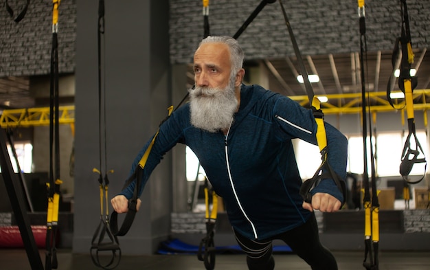 Barbudo deportista senior entrenamiento con bandas de resistencia trx en el gimnasio.