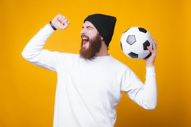 Barbudo hombre alegre está gritando y celebrando, sosteniendo un balón de fútbol en la pared amarilla.