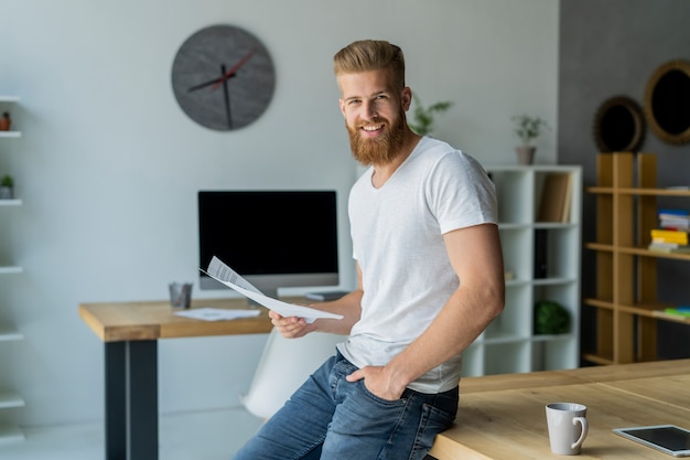 Barbudo jovem empresário trabalhando em um escritório moderno. Homem vestindo camiseta branca e fazendo anotações nos documentos.