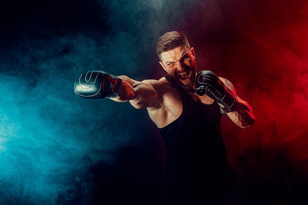 Barbudo tatuado desportista muay thai boxer em camiseta preta e luvas de boxe lutando em um fundo escuro com fumaça. conceito de esporte.