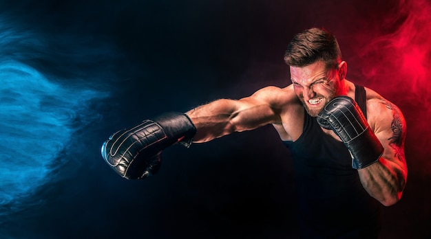 Barbudo tatuado desportista muay thai boxer em camiseta preta e luvas de boxe lutando em um fundo escuro com fumaça. conceito de esporte.