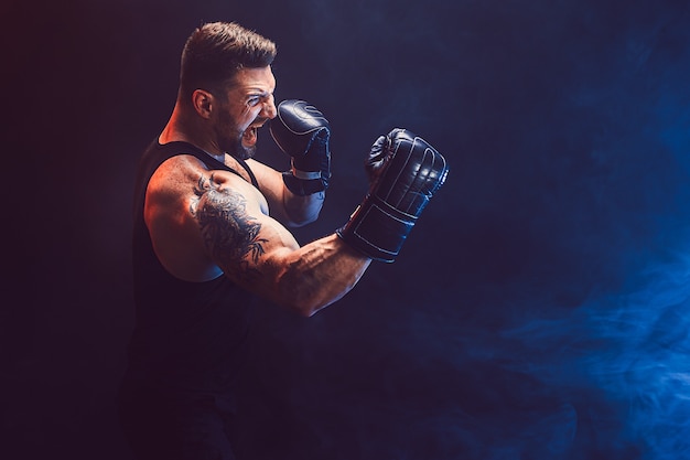 Barbudo tatuado desportista muay thai boxer em camiseta preta e luvas de boxe lutando na parede escura com fumaça. Conceito de esporte.