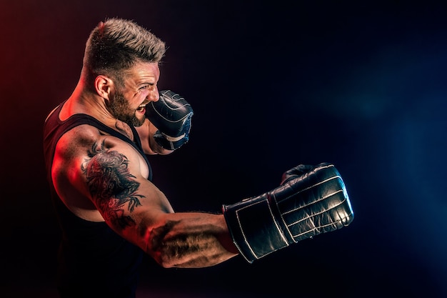 Barbudo tatuado desportista muay thai boxer em camiseta preta e luvas de boxe lutando na parede escura com fumaça. Conceito de esporte.