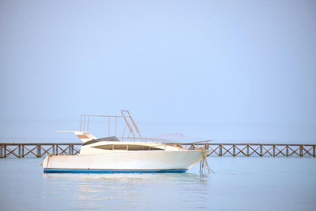 Barco a motor branco flutua na água do oceano na ponte longa do cais sob a vista do céu azul brilhante da praia de areia Conceito de viagem de verão