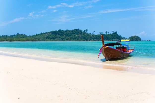 Barco de madeira em uma praia de areia branca, mar azul com ilhas no fundo, praia tropical na Tailândia
