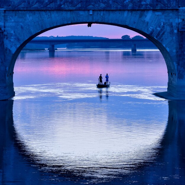 Barco de pesca sob o arco de uma ponte em um pôr do sol colorido.