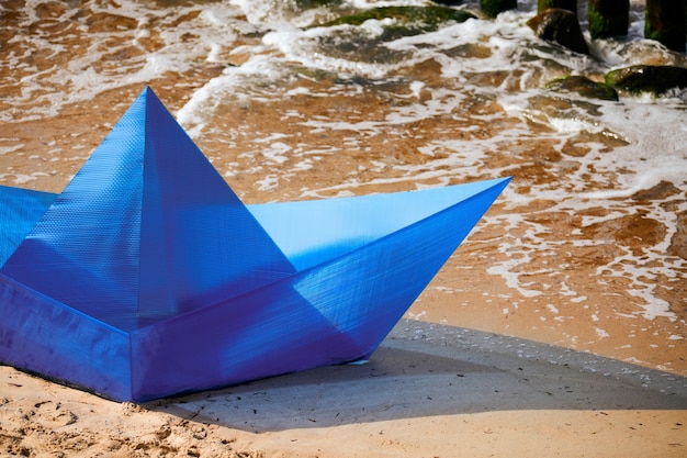 Barco de papel azul de origami en la playa de arena para el diseño de concepto, hermoso barco de papel en la costa. Objeto de arte moderno, primer plano, fondo del océano. Diversión de verano al aire libre, viajes, concepto de vacaciones de verano