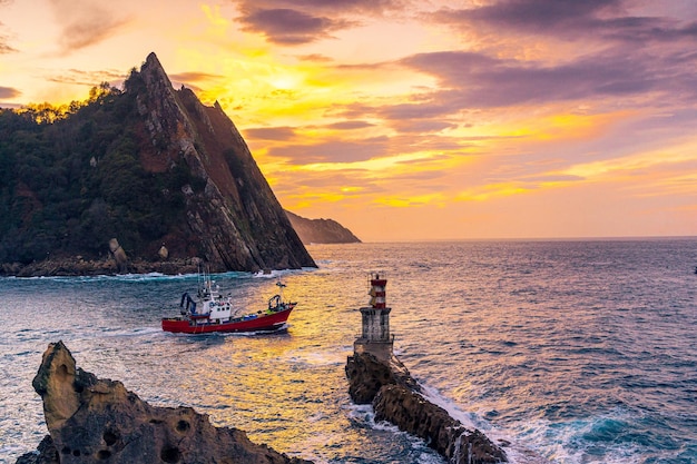 Un barco saliendo en el atardecer naranja del faro de Pasajes San Juan