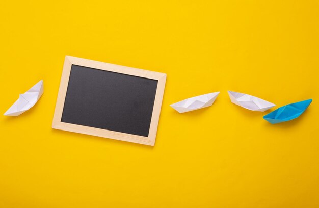 Barcos de papel y mini pizarra en blanco con espacio de copia sobre un fondo amarillo Liderazgo empresarial o concepto de viaje Vista superior Plano