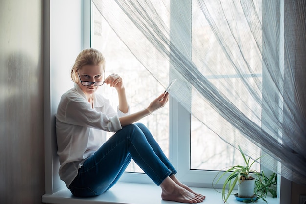 Barfüßige Frau mit Brille und weißem Hemd, die auf der Fensterbank sitzt und das Smartphone in der Hand hält Süße Blondine, die sich die Kamera in der Nähe ansieht