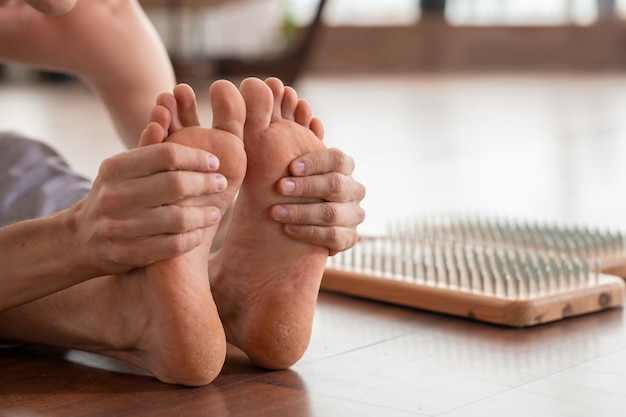 Barfuß Mann sitzt auf Holzboden mit Yoga-Therapie-Pads mit metallischen Nägeln in der Nähe und hält seine Füße während einer der Übungen in den Händen