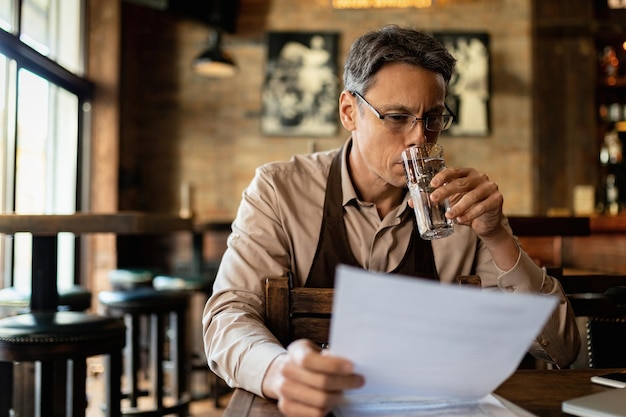 Barista adulto medio leyendo informes mientras toma un vaso de agua en un pub