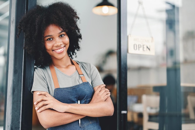 Barista afroamericano sonriente con delantal de pie en la puerta de un café