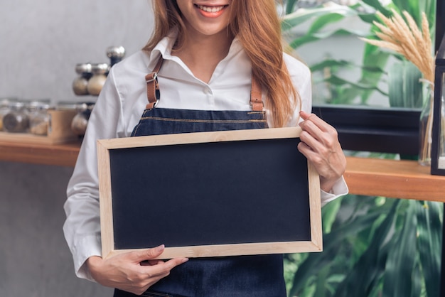 Barista asiática joven que sostiene el menú de la pizarra en blanco y que sonríe en el fondo contrario del café