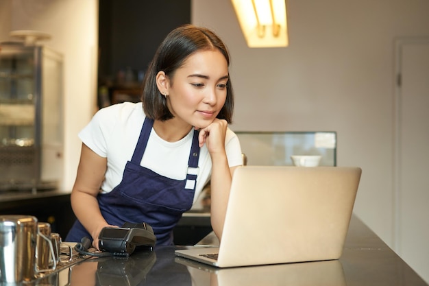 Una barista asiática sonriente que trabaja en un café sirviendo a un cliente mirando una computadora portátil mientras usa un lector de tarjetas