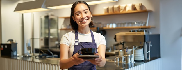 Barista asiática sorridente fazendo café segurando uma xícara usando avental uniforme de café em pé perto