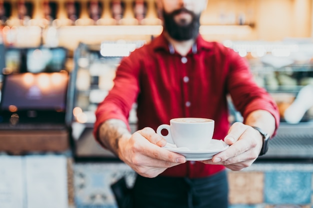 Barista barbudo hermoso joven en camisa roja que trabaja en la barra de café moderna.