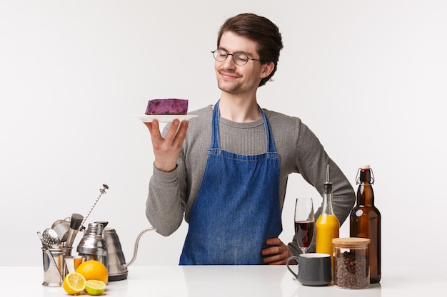 Barista, Cafe Worker und Barkeeper-Konzept. Porträt des erfreuten lächelnden, glücklichen jungen männlichen Kleinunternehmers, der ein Stück Kuchen auf Teller mit zufriedenem Lächeln hält, stehen nahe der Bartheke