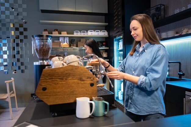 Barista caucasiana concentrada a operar a máquina de café ao lado da sua colega asiática satisfeita.