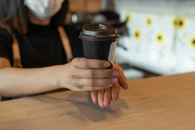 Barista de mulher amigável usando máscara protectora à espera de servir café preto gelo ao cliente no café café, restaurante café, mente de serviço, pequeno empresário, conceito de comida e bebida