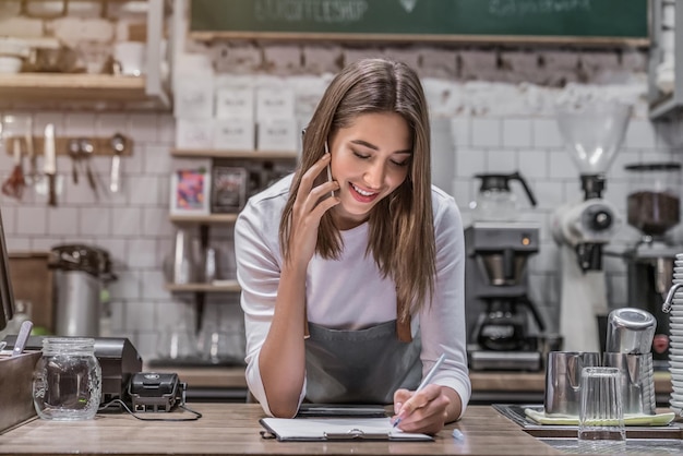 Barista de mulher feliz trabalhando sorrindo e falando no smartphone aceitando ordem de escrita