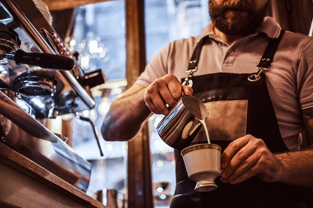 Barista en delantal haciendo un capuchino, sirviendo leche en una taza en un restaurante o cafetería