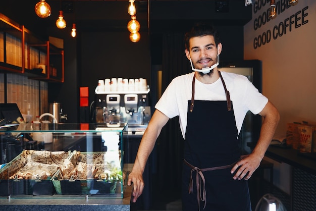 Barista en delantal con mascarilla de pie en el bar de la cafetería.