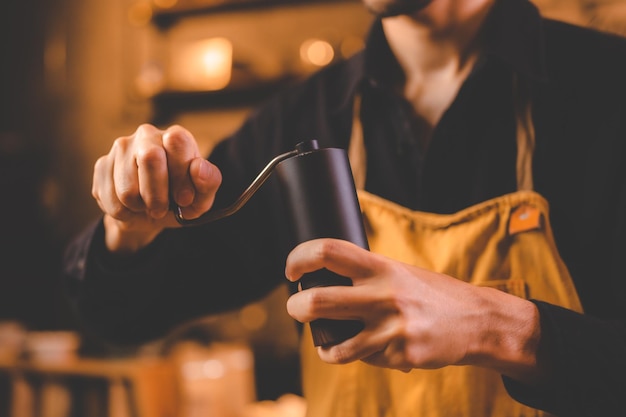 Barista, der eine Handmühle hält, um Getränke zuzubereiten, indem er geröstete Kaffeebohnen für Koffeingetränke in der Küche, Café, Hintergrund, morgendliche Frühstücksmühlenzubereitung mit Espressoausrüstung in braunem Ton mahlt