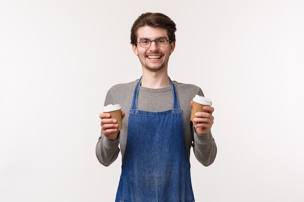 Barista, ein Porträt eines unbeschwerten, lächelnden, freundlichen männlichen Angestellten in der Schürze, machte köstlichen Kaffee für die Kunden, hielt zwei Tassen zum Mitnehmen und lachte und lud ein, den besten Cappuccino der Stadt zu probieren