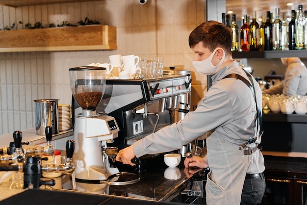 Un barista enmascarado prepara un exquisito y delicioso café en el bar de una cafetería. El trabajo de los restaurantes y cafeterías durante la pandemia.