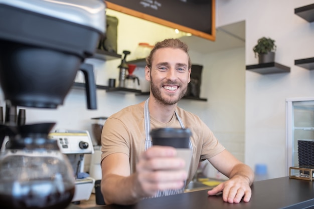 Barista. Freudiger bärtiger junger männlicher Barista an der Theke des Cafés, das höflich ein Glas Kaffee heraushält