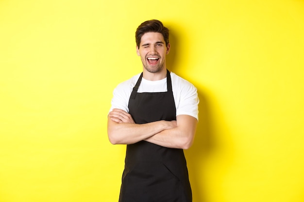 Barista guapo en delantal negro guiñando un ojo, vestido con uniforme de delantal negro, de pie sobre la pared amarilla