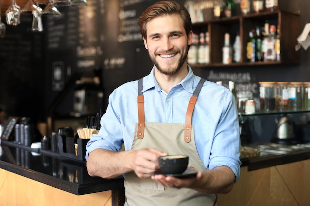 Barista guapo que ofrece una taza de café a la cámara en la cafetería.