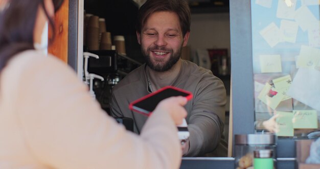 Foto barista-mann macht kaffee zum mitnehmen für eine kundin, die per kontaktlosem mobiltelefon an das kreditkartensystem zahlt