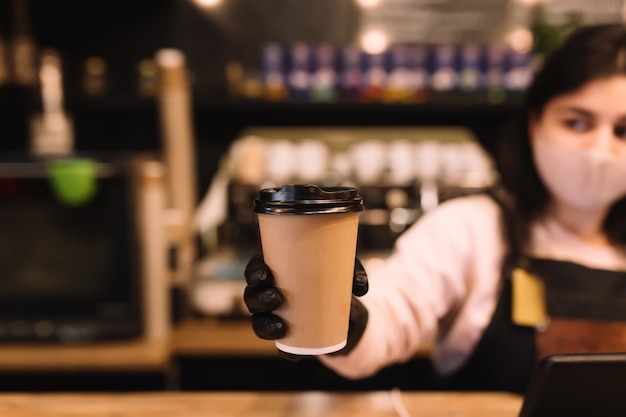 Barista en mascarilla protectora y guantes negros da una taza de café en la cafetería.