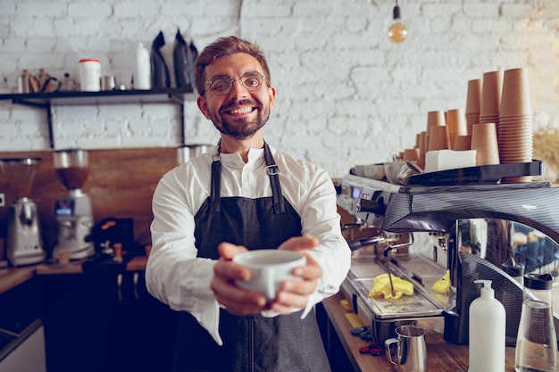 Barista masculino alegre que ofrece una taza de café en la cafetería
