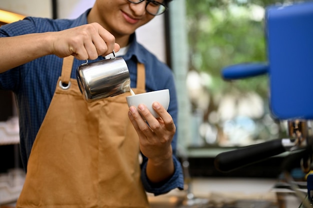 Barista masculino asiático talentoso que trabaja en la cafetería que vierte leche al vapor en una taza