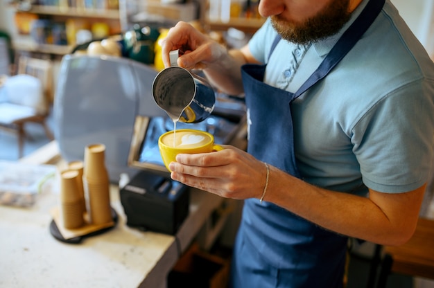 Barista masculino en delantal vierte espuma en el café en la cafetería. El hombre hace espresso fresco en la cafetería, el camarero en el mostrador de bar