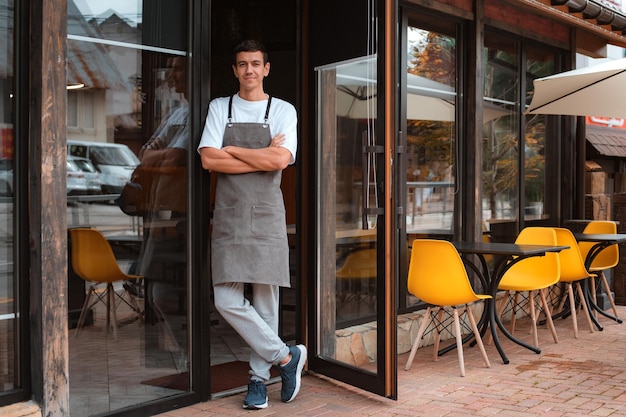 Barista ou garçom de café ou dono de cafeteria contra gesto de entrada convidando você a visitar um cara sorridente de avental ao ar livre, orgulhoso de seu pequeno negócio local