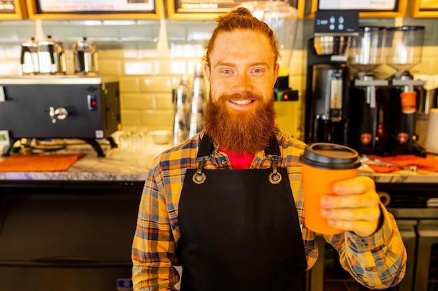 Barista profesional joven barbudo de jengibre pelirrojo con delantal negro trabajando en una cafetería