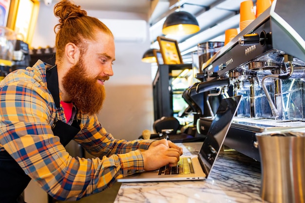 Barista profesional joven barbudo de jengibre pelirrojo con delantal negro trabajando en una cafetería