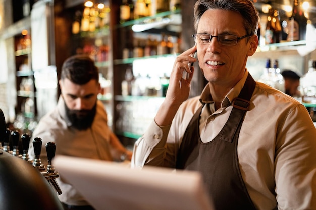 Foto barista sorridente fazendo uma ligação enquanto lê a lista de inventário em um bar